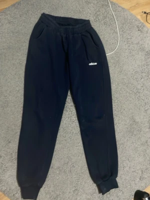 Mörkblå mjukisbyxor från Adidas - Säljer ett par mörkblå mjukisbyxor från Adidas med broderad logga på benet. Byxorna har resår i midjan och vid bensluten samt två sidofickor. Perfekta för chill dagar eller när du vill ha det extra bekvämt.