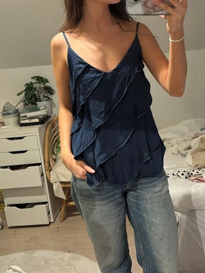 Mörkblå linnetopp med volang - Snygg mörkblå linnetopp med tunna axelband och v-ringning. Toppen har flera lager volangdetaljer framtill som ger en cool och unik look. Perfekt att styla med jeans eller kjol för en trendig outfit.