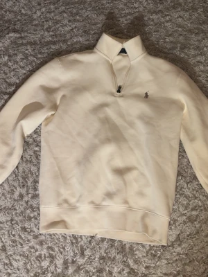 Vit halvzip tröja från Polo Ralph Lauren - Snygg vit sweatshirt med halv dragkedja från Polo Ralph Lauren. Tröjan har hög krage och den klassiska broderade loggan på bröstet. Tillverkad i mjukt material och har ribbade muddar vid ärmslut och nederkant. Perfekt för en clean och stilren look.