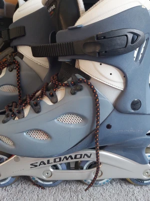 Salomon inlines grå och vit - Salomon inlines med grått och vitt hårdplastskal, svart och orange snörning samt svart spänne. Metallram med synliga hjul och ventilerade partier på sidorna. Märkt Human Print Auto Fit på plösen.