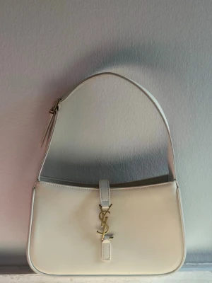 Vit axelremsväska från Saint Laurent - Stilren vit handväska från Saint Laurent i slätt läder med axelrem och guldfärgat YSL-monogram framtill. Väskan har en klassisk, rektangulär form och stängs med ett lock och magnetknäppning. Perfekt för dig som gillar lyxiga accessoarer.