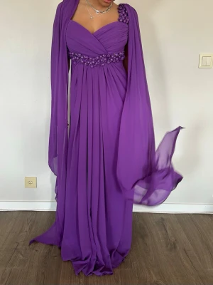 Lila balklänning💜 - Jätte fin lila långklänning med omlott över bysten och v-ringad urringning. Klänningen har pärldetaljer i midjan och på axelbanden samt ett matchande sjal-liknande tyg. Tillverkad i ett lätt, flytande material som ger ett drömmigt intryck. Perfekt för bal eller student💜