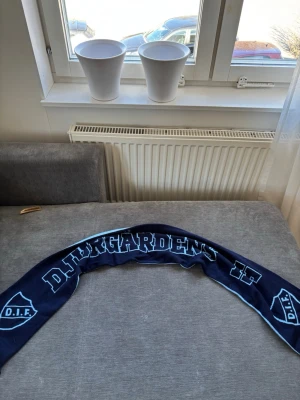 Djurgårdens IF halsduk blå - Mörkblå halsduk med ljusblå text och DIF-logga på båda sidor. Perfekt för dig som vill visa ditt stöd för Djurgårdens IF. Tjockt tyg som håller dig varm under matcherna. Klassisk supporterdesign.