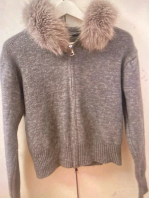 Grå ziptröja med fuskpäls - Mysig grå ziptröja med fluffig fuskpäls runt kragen. Tröjan har långa ärmar, ribbade muddar och dragkedja framtill. Perfekt för dig som vill ha en trendig och bekväm look med lite extra detaljer.