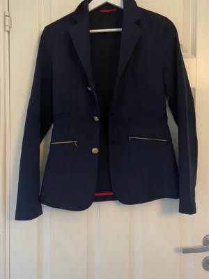 Mörkblå kavaj med guldknappar - Stilren mörkblå kavaj (till ridning men funkar även till annat) med två guldknappar framtill och dekorativa dragkedjor vid fickorna. Kavajen har klassisk krage, långa ärmar och röda detaljer baktill. Materialet ser ut att vara en mjuk bomullsblandning. Perfekt för dig som vill ha en snygg och modern look. (Inköpspris 1000kr)