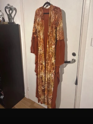 Kurdiska klänningen xs/s - Säljer en lång, brun morgonrock med unikt mönster i guldiga och beige toner. Morgonrocken har lång ärm och omlottmodell med bälte i midjan. Perfekt för att chilla hemma i stil.