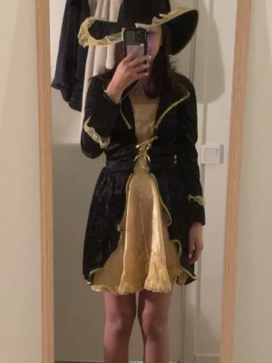 Svart och gul piratutkläddnad med hatt - Säljer en svart och gul piratutkläddnad  med matchande stor hatt. Klänningen har guldfärgade detaljer och volanger, samt långa ärmar med dekorativa kanter. Perfekt för maskerad eller cosplay och ger en magisk vibe. köpte denna till halloween men ändra till en annan kostym. passar bra för utkläddnadsfester och teman. 