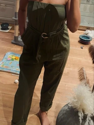 Olivgrön jumpsuit från Zara - Säljer en olivgrön jumpsuit från Zara i storlek S. Axelbandslös modell med avtagbart midjebälte och fickor. Tillverkad i ett lätt tyg som känns skönt mot huden. Perfekt för dig som vill ha en trendig och bekväm look.