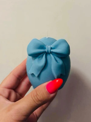 Blå dekorativt ljus med rosett - Säljer ett blått dekorljus i form av ett ägg med en stor, detaljerad rosett på framsidan. Ljuset är tillverkat av vax och levereras i en ljusrosa presentförpackning med fönster. Perfekt som inredningsdetalj eller present.