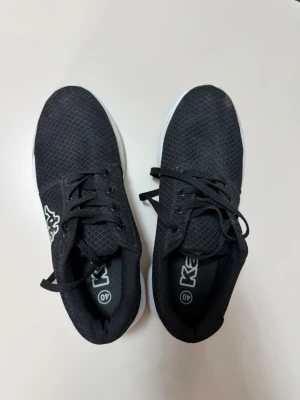 Svarta sneakers från Kappa - Svarta sneakers från Kappa med vit sula och klassisk låg profil. Ovandelen är i meshmaterial för bra andningsförmåga och skorna har svarta skosnören. Perfekta för en sportig och avslappnad stil.