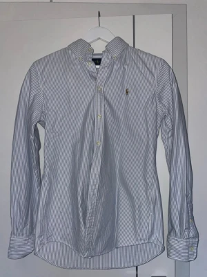 Randig skjorta med knappkrage - Säljer en klassisk vit skjorta med tunna blå ränder och knappkrage. Skjortan har långa ärmar, knappar framtill och en liten broderad logga på bröstet. Perfekt till jeans eller chinos för en clean och stilren look.