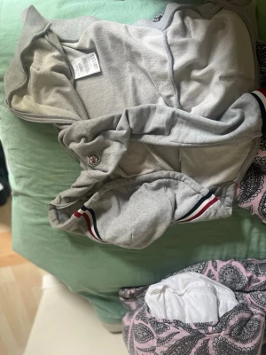 Grå hoodie från Moncler med huva - Säljer en grå hoodie från Moncler med klassisk huva och snygga ränder i rött, vitt och blått vid ärmsluten. Hoodien har en broderad Moncler-logga på ärmen och är tillverkad i mjukt bomullsmaterial. Perfekt för en avslappnad och trendig look.