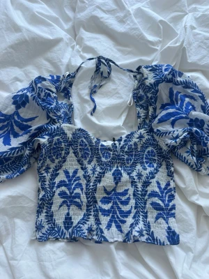Blåvit blommig croppad blus - Trendig croppad blus i blått och vitt med blommigt mönster. Puffiga korta ärmar och knyt i nacken ger en romantisk vibe. Tillverkad i ett lätt och luftigt material, perfekt för varma dagar. Snygg till både jeans och kjol!