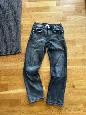 Grå raka jeans med fem fickor - Säljer ett par grå jeans med rak passform och klassisk femficksdesign. Jeansen har en något tvättad look och stängs med dragkedja och knapp. Materialet är denim i bomull och de har bälteshällor i midjan.