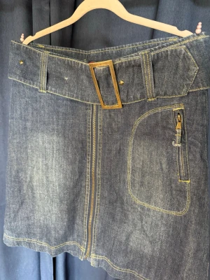 Jeanskjol med bredt bälte och ficka - Snygg jeanskjol i mörkblå denim med bredt bälte och stor guldfärgad spänne. Kjolen har en stor ficka med dragkedja och dekorativa sömmar i gult. Perfekt för en trendig och avslappnad stil.