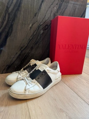 Valentino Garavani vita sneakers - Snygga vita sneakers från Valentino Garavani med en bred svart läderdetalj över mitten. Klassisk låg modell med vita snören och diskret logga på plösen. Skorna är tillverkade i läder och har en stilren design som sticker ut.