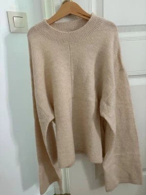 Beige oversized stickad tröja - Mysig beige stickad tröja med oversized passform och bred ribbad rund halsringning. Tröjan har långa ärmar och är tillverkad i ett mjukt material som ger en avslappnad look. Perfekt för lager på lager och chill dagar.