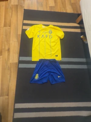 Gul Nike fotbollströja och blå shorts - Säljer ett matchande set med gul Nike Dri-Fit fotbollströja och blå shorts. Tröjan har tryck framtill och tunna blå linjer, medan shortsen har klubbmärke och Nike-logga. Perfekt för träning eller match. Materialet är lätt och andas bra.
