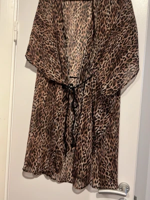 Leopardmönstrad strandkimono - Snygg och luftig strandkimono med leopardmönster i bruna och svarta toner. Modellen är öppen framtill och har ett knytband i midjan. Perfekt att slänga över bikinin på stranden. Tillverkad i ett tunt, transparent material som känns lätt och bekvämt.
