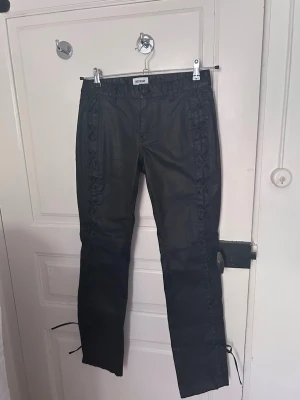 Svarta skinnbyxor med snörning - Säljer ett par svarta skinnbyxor med snygga snörningsdetaljer längs sidorna. Byxorna har raka ben och hällor för skärp. Storlek 34/XS. Low/mid waist :)