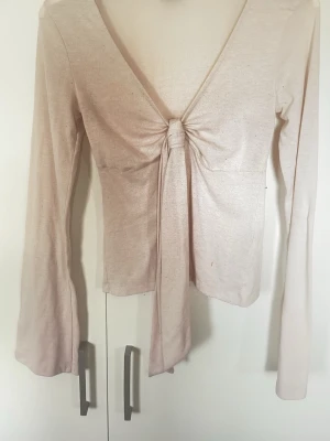 Ljusbeige topp med knyt framtill - Rosa topp med lång ärm och djup v-ringning. Toppen har ett snyggt knyt framtill. Perfekt att styla med både jeans och kjol för en söt outfit.