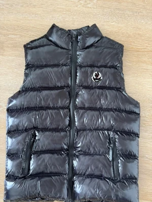 Svart glansig dunväst med dragkedja - Snygg svart dunväst med glansig finish och quiltad design. Västen har hög krage, två dragkedjeförsedda fickor och ett emblem på bröstet. Perfekt för lager-på-lager och håller dig varm under kyliga dagar.