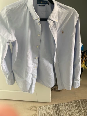 Ljusblå skjorta från Ralph Lauren - Klassisk ljusblå skjorta från Ralph Lauren med button-down krage och broderad logga på bröstet. Skjortan har långa ärmar, vita knappar och är tillverkad i bomull. Perfekt för en stilren och avslappnad look. Skjortan är oanvänd och i bra form!