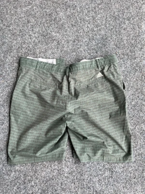 Gröna randiga shorts från Adidas - Snygga gröna shorts från Adidas med diskret randigt mönster. Klassisk passform med bälteshällor och fickor både fram och bak. Perfekta för sommardagar och avslappnade tillfällen.