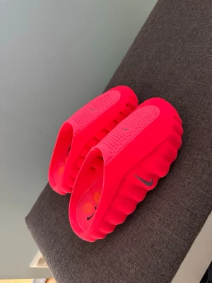 Nike mind 001 - Säljer ett par neonrosa tofflor från Nike med vågig sula och ventilerande hål på ovansidan. Tofflorna har en chunky design och är tillverkade i ett mjukt, flexibelt gummimaterial. Perfekta för dig som vill sticka ut med färg och komfort.