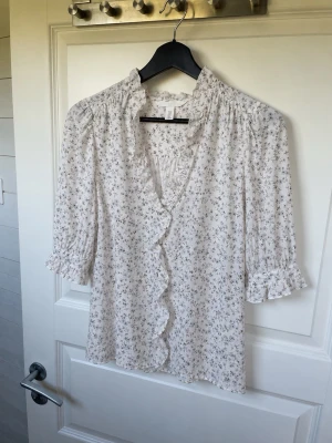 Blommig blus med volang från H&M - Söt blommig blus från H&M i vitt med små grå och blå blommor. Blusen har volangkant längs knäppningen, rynkad krage och trekvartsärm med volang vid ärmslut. Perfekt för en romantisk och trendig look.