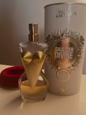 Jean Paul Gaultier Divine parfym - Unik parfymflaska formad som en kvinnokropp med guldfärgade detaljer och konformad byst. Flaskan har en transparent glasdesign och kommer med en elegant vit och guldig förpackning. Perfekt för dig som gillar lyxiga och iögonfallande dofter.