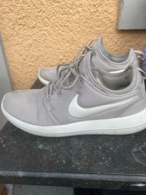 Grå Nike sneakers med vit sula - Snygga grå Nike sneakers med vit sula och klassisk vit swoosh på sidan. Ovandelen är i textil och skon har låga snörningar. Sulan har ett rutmönster undertill med gröna detaljer. Perfekta för en avslappnad streetstyle.