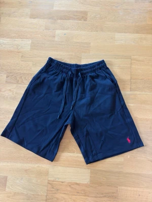 Marinblå shorts från Polo Ralph Lauren - Snygga marinblå shorts från Polo Ralph Lauren med röd broderad logga på benet. Shortsen har elastisk midja med snörning och är perfekta för sommaren. Tillverkade i mjuk bomull för en skön känsla.