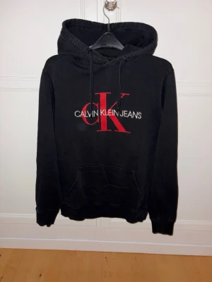 Svart hoodie Calvin Klein - Svart hoodie med stor röd CK-logga och vit text på bröstet. Klassisk känguruficka framtill och justerbar huva med snören. Mjuk bomullskänsla och ribbade muddar vid ärmslut och nederkant. Perfekt för en avslappnad och stilren look.