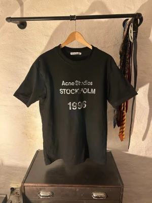 Svart Acne Studios t-shirt - Säljer en svart t-shirt från Acne Studios med tryck i vitt och silver på bröstet där det står 'Acne Studios STOCKHOLM 1996'. Klassisk rund hals och korta ärmar. Tillverkad i mjuk bomull för en skön känsla.