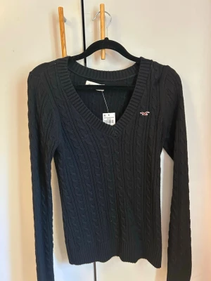 Svart kabelstickad tröja Hollister - helt ny med lapp kvar! Kabelstickad svart tröja från Hollister med V-ringning och lång ärm. Tröjan har ribbade muddar och en liten broderad logga på bröstet. 