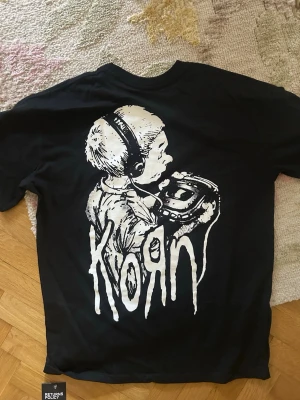 Svart Korn band-tshirt med tryck - Svart t-shirt med stort tryck av en tecknad figur och Korn-loggan på ryggen samt texten 'Follow the Leader' på bröstet. Klassisk passform och rund hals. Perfekt för dig som gillar bandmerch och vill sticka ut med en cool design.
