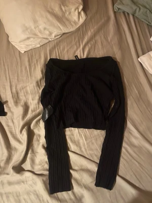 Svart ribbad offshoulder topp - Snygg svart ribbad topp med offshoulder-design och långa ärmar. Croppad modell som passar perfekt till höga jeans eller kjol. Trendig och enkel att styla för en cool look.