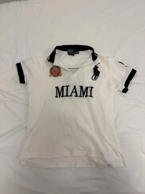 Vit pikéskjorta Polo Ralph Lauren - Vit pikéskjorta från Polo Ralph Lauren med svart krage och svarta detaljer. Stor MIAMI-text framtill, broderad logga och patch på bröstet. Klassisk och sportig look med korta ärmar och knappar vid halsen.