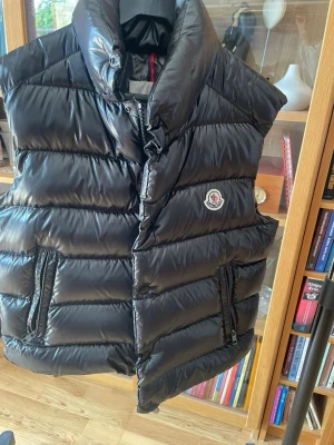 Svart dunväst från Moncler - Snygg svart dunväst från Moncler med glansig finish och hög krage. Västen har två dragkedjeförsedda fickor och Monclers klassiska logotyp på bröstet. Perfekt för lager-på-lager och håller dig varm under kyliga dagar.