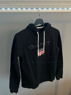 Svart hoodie från GANT med tryck - Svart hoodie från GANT med vit dragsko i huvan och stort GANT-tryck på bröstet tillsammans med ett broderat emblem i rött, vitt och blått. Klassisk passform och mjukt material, perfekt för en avslappnad stil.