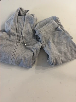 Grått loungewear-set från Gina Tricot - Mysigt grått loungewear-set från Gina Tricot med dragkedja och huva på tröjan samt matchande byxor med resår i midjan. Perfekt för chill dagar hemma. Materialet är mjukt och skönt mot huden. Storlek 146/152 i tröjan och 134/140 i byxorna. 