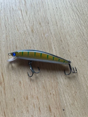 Fiskedrag i grönt och gult - Säljer ett fiskedrag med grön och gul randig kropp, blått öga och två trekrokar. Draget har en smal, avlång form och en liten plastsked framtill. Perfekt för fiske efter rovfiskar.
