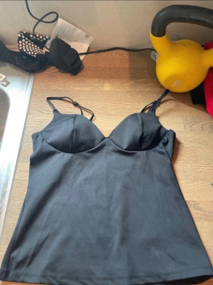 Svart topp från Gina Tricot - Snygg svart topp från Gina Tricot med tunna justerbara axelband och formpressade kupor. Tillverkad i ett mjukt och stretchigt material som sitter skönt mot kroppen. Perfekt att styla under en skjorta eller kavaj.