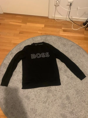 Svart sweatshirt från BOSS - Snygg svart sweatshirt från BOSS med vit broderad logga på bröstet. Tröjan har rund halsringning och långa ärmar. Perfekt för en avslappnad och trendig stil.