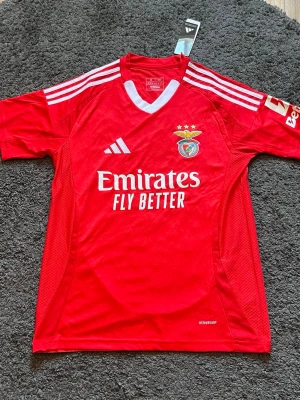 Benfica matchtröja  - Säljer en röd Benfica matchtröja från Adidas i storlek M. Tröjan har vita detaljer, klubbmärke på bröstet, Adidas-logga och sponsortryck 'Emirates Fly Better' framtill. Tillverkad i lätt och ventilerande material med slim fit passform.