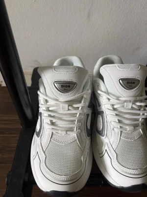 Vita Dior B30 sneakers - Snygga vita Dior B30 sneakers med mesh och läderdetaljer. Skorna har en chunky sula, vita skosnören och silverfärgad logga på plösen. Stora D-detaljer på sidan i grått och svart ger en modern vibe. Perfekta för dig som vill sticka ut med exklusiva sneakers. Dem är storlek 40 men lite små så skulle säga typ 39,5,skriv om ni är intresserade!!