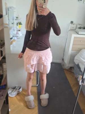 Rosa rutiga volangshorts - Supersöta rosa shorts med rutigt mönster och volangdetaljer. Högmidjade med smock i midjan för en bekväm passform. Perfekta för sommarens varma dagar och ger en gullig och trendig look.