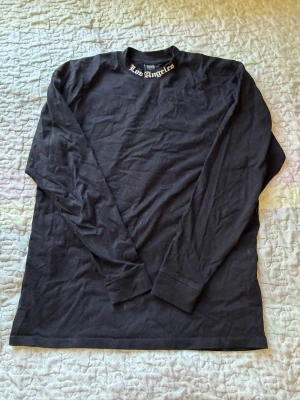 Svart långärmad topp Black Squad - Svart långärmad topp från Black Squad med texten 'Los Angeles' tryckt vid halsen och ryggen, samt längst ner på ryggen. Tight fit och tillverkad i mjuk bomull. Perfekt för en avslappnad och cool stil.