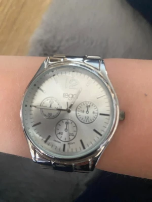 Silverfärgad klocka från Regal - Stilren klocka från Regal i silverfärg med metallarmband och rund urtavla. Urtavlan har tre dekorativa små ur och tydliga markeringar. Perfekt accessoar för dig som gillar en klassisk look.